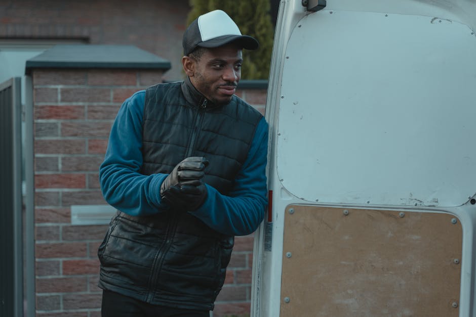 A man wearing a black and grey baseball cap, black gloves, a dark padded vest, and a blue long-sleeved shirt is standing outdoors next to an open small white van, which is positioned near a residential property with a brick wall and greenery in the background. The man is in the process of loading or unloading items, possibly during a home relocation or furniture transport, as he holds a cardboard box wrapped in plastic wrap. Inside the van, visible packing materials such as cardboard and plastic wrap can be seen, along with a few other boxes and packing supplies stacked inside. The loading area appears well-lit with natural daylight, and the scene depicts the logistics of packing and moving with the man actively engaged in the loading process, demonstrating the typical handling involved in house removals or professional moving services associated with Man with Van Maida Vale.