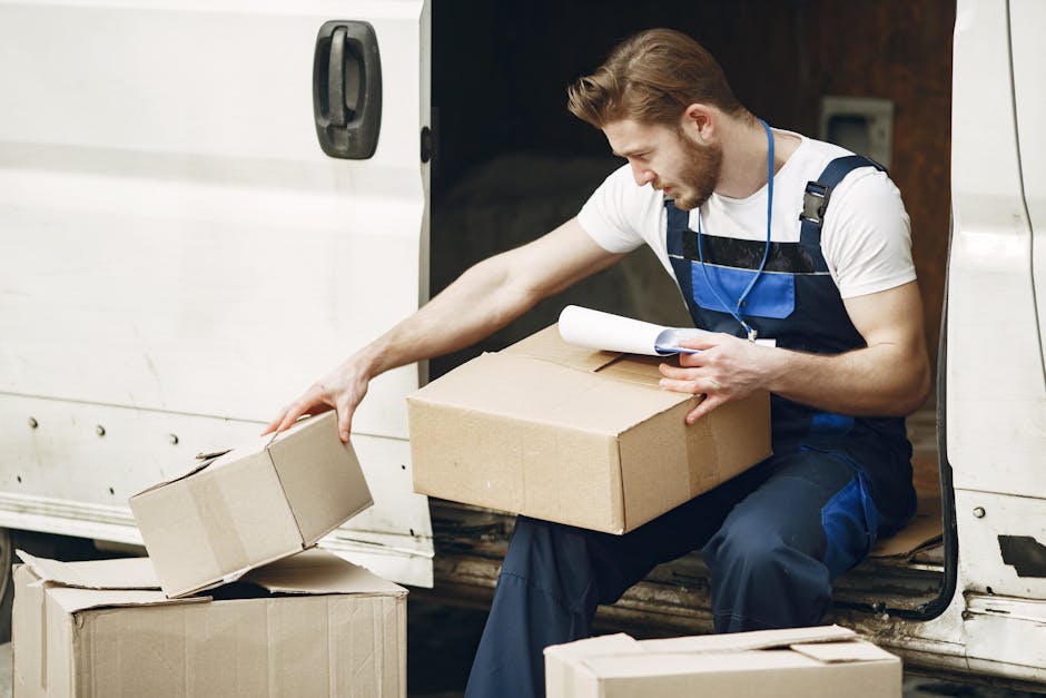 A man with light brown hair and a beard, dressed in a white t-shirt and navy blue work overalls, is sitting on the edge of a white van with its side door open. He is handling cardboard boxes, one in each hand, with additional boxes placed on the ground nearby. The man appears to be examining a set of plans or a survey document held in his left hand. The scene takes place on a pavement area, and the van's interior, partially visible, is used for home relocation and furniture transport. Safety equipment such as a harness is worn around his neck, indicating careful handling during packing and loading processes, which are essential elements of professional removals provided by Man with Van Maida Vale. The lighting is natural, highlighting the packaging materials, including cardboard boxes, and the focused activity of loading or preparing for a move as part of a house removal service. This image visually supports the themes of packing, moving logistics, and home relocation services.