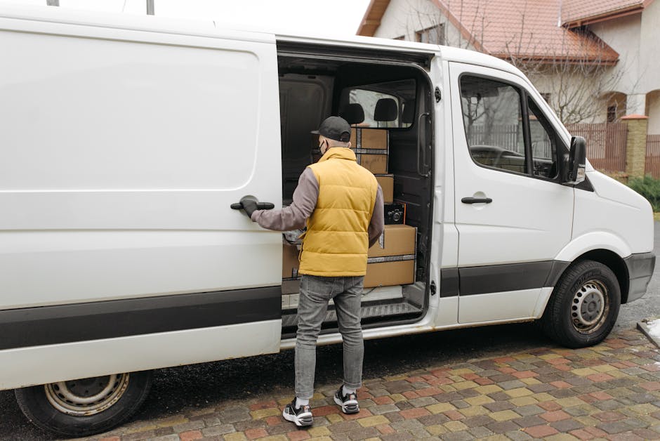 A man wearing a black and grey baseball cap, black gloves, a dark padded vest, and a blue long-sleeved shirt is standing outdoors next to an open small white van, which is positioned near a residential property with a brick wall and greenery in the background. The man is in the process of loading or unloading items, possibly during a home relocation or furniture transport, as he holds a cardboard box wrapped in plastic wrap. Inside the van, visible packing materials such as cardboard and plastic wrap can be seen, along with a few other boxes and packing supplies stacked inside. The loading area appears well-lit with natural daylight, and the scene depicts the logistics of packing and moving with the man actively engaged in the loading process, demonstrating the typical handling involved in house removals or professional moving services associated with Man with Van Maida Vale.