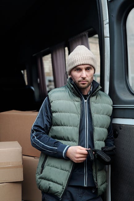 A man with light skin and a serious expression stands beside a moving van during a house relocation. He is wearing a beige knitted beanie, a grey padded vest over a navy blue and grey jacket, and dark gloves. In his hands, he holds a black strap used for securing furniture during transport. The open sliding door of the van reveals cardboard moving boxes stacked inside, with some boxes partially visible behind him. The interior of the van appears dark with a curtain covering the window. The setting suggests the loading process of furniture and packed belongings as part of a professional removals service provided by Man with Van Maida Vale, supporting house and office moves in the Maida Vale area.