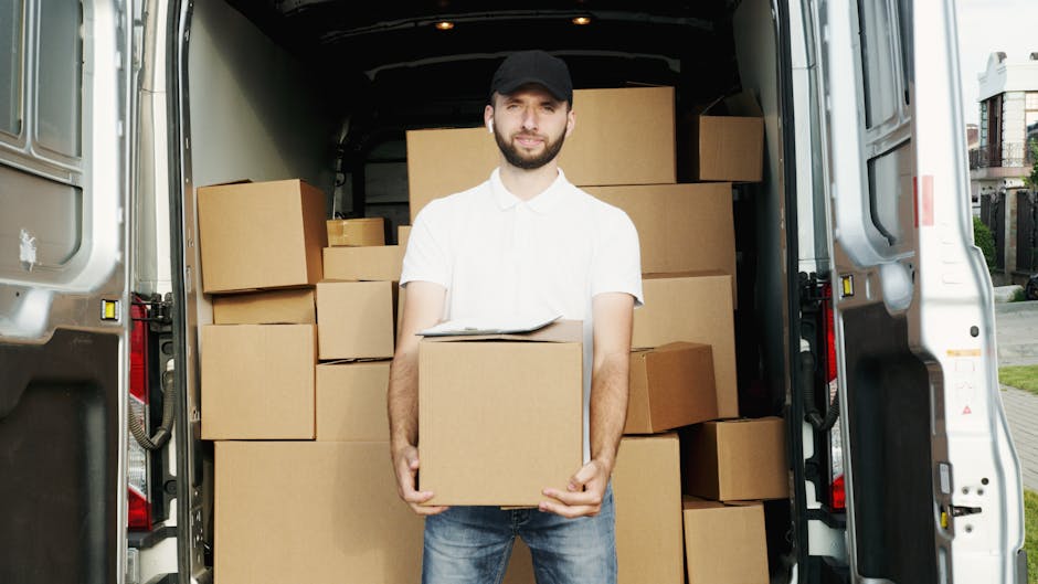 A man standing at the open back of a white van loaded with several cardboard boxes, some stacked on top of each other inside the vehicle. He is holding a medium-sized cardboard box with both hands, and there is a clipboard or paper resting on top of it. The boxes are wrapped in brown packing paper, and some are secured with plastic wrap. The scene takes place in an outdoor residential area with pavement, and part of a house or building is visible in the background. The lighting suggests daytime, and the man is dressed in a white short-sleeved shirt, blue jeans, and a black cap. The van's interior appears clean and organized, indicative of a professional house or office relocation service. This image highlights the loading process during furniture transport and packing for a move, consistent with services provided by Man with Van Maida Vale, specializing in removals and home relocation.