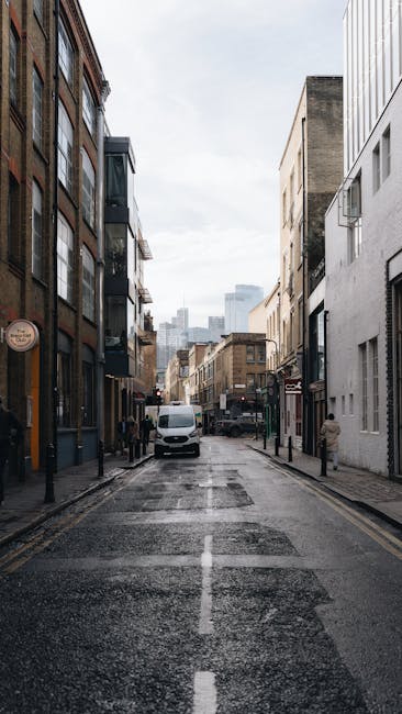 A narrow urban street during daytime, with multi-story buildings on both sides featuring a mix of brick, glass, and concrete facades. The street surface is wet, reflecting the overcast sky above. Seve