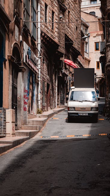 A narrow urban street during daytime, with multi-story buildings on both sides featuring a mix of brick, glass, and concrete facades. The street surface is wet, reflecting the overcast sky above. Seve