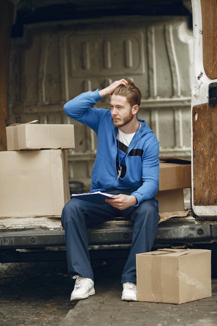 A young male mover with light skin, wearing a blue tracksuit and white sneakers, is sitting on the edge of a van’s open cargo area during a home relocation process. He is holding a clipboard in his left hand and scratching his head with his right, appearing to be in thought or planning. Inside the van, there are several cardboard boxes of various sizes, some sealed with tape, positioned on the floor and stacked on one side. The interior of the van is lined with a grey, ribbed metal surface, and a wooden panel is visible on the right side near the door. Outside, the van is parked on a paved area, with additional cardboard boxes placed on the ground nearby, some partially visible. Straps, blankets, and packing materials are not visible in the image. This scene reflects the loading or unloading phase of a furniture transport and packing service offered by [COMPANY_NAME], emphasizing careful planning and organisation during the shifting process as part of house removals in Maida Vale.