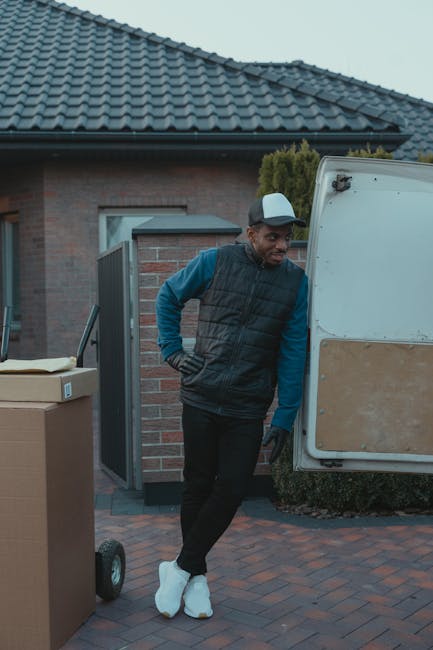 A man dressed in a dark padded vest, blue long-sleeve shirt, black trousers, and white sneakers stands next to a white moving van with a wooden interior panel on the side, parked on a brick driveway outside a residential property. He is wearing a light-colored cap and has his hands in his pockets, observing the loading area. Nearby, there are several cardboard boxes and a flatbed trolley, likely used for transporting household items during a house relocation. The background features part of a modern brick house with a grey tiled roof, a window, and a hedge. The lighting suggests an overcast day, capturing the typical scene of furniture transport and packing preparations associated with removals and furniture transport services offered by [COMPANY_NAME]. This image visually reflects the process of home relocation, including loading and packing activities for a move in Maida Vale, aligned with professional removals services.