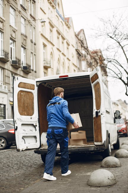 A young male mover with light skin, wearing a blue tracksuit and white sneakers, is sitting on the edge of a van’s open cargo area during a home relocation process. He is holding a clipboard in his left hand and scratching his head with his right, appearing to be in thought or planning. Inside the van, there are several cardboard boxes of various sizes, some sealed with tape, positioned on the floor and stacked on one side. The interior of the van is lined with a grey, ribbed metal surface, and a wooden panel is visible on the right side near the door. Outside, the van is parked on a paved area, with additional cardboard boxes placed on the ground nearby, some partially visible. Straps, blankets, and packing materials are not visible in the image. This scene reflects the loading or unloading phase of a furniture transport and packing service offered by [COMPANY_NAME], emphasizing careful planning and organisation during the shifting process as part of house removals in Maida Vale.