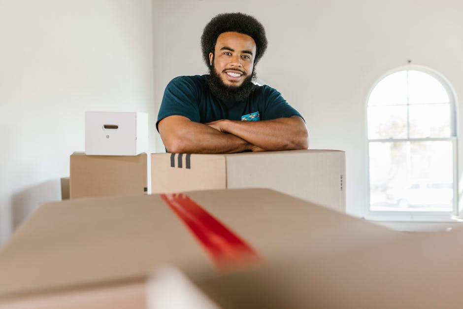 A smiling man with curly black hair and a beard, wearing a dark blue T-shirt, is standing behind a stack of cardboard boxes inside a well-lit room with cream walls and a large, arched window. The boxes, some sealed with red and black packing tape, are arranged on a surface, with smaller boxes positioned on top of larger ones. The background shows part of the room's interior, including a window allowing natural light to fill the space. This scene depicts the unpacking or packing process involved in home relocation or furniture transport, with the man from Man with Van Maida Vale preparing items for a removal service, emphasising careful handling and organisation of household goods for an efficient move.