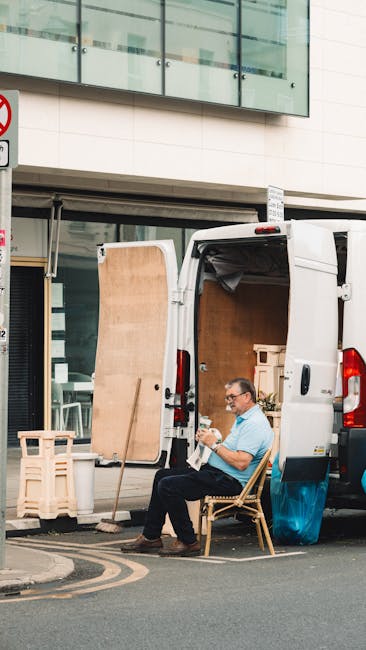 A man with curly hair wearing a blue T-shirt and dark jeans sits on the wooden floor of a bright room with large arched windows, which reveal a street view with parked cars and trees outside. He is surrounded by several cardboard moving boxes, some sealed with red tape, indicating they are prepared for a house relocation. The room is illuminated by natural light streaming through the windows, casting soft shadows in the space. In the corner, a tall potted plant adds greenery to the scene. The boxes are stacked and positioned near the man, suggesting they are part of a packing and moving process. The setting appears to be inside a residential property, with a focus on interim storage during the packing and loading stages of the relocation, a service offered by Man with Van Maida Vale.