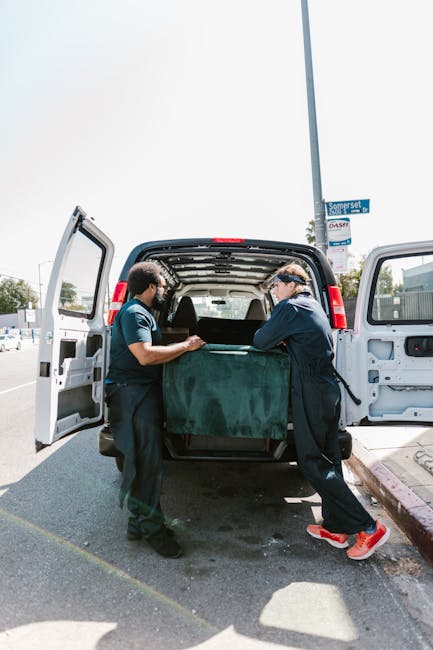 Two men wearing dark uniforms are loading a large, dark green sofa into the back of a white moving van parked on a paved street. The van's rear doors are open, revealing the interior with more packing materials and possibly other furniture. One man is holding the sofa steady while the other pushes it into the vehicle, both focused on the loading process. The background includes a lamppost, a street sign reading 'Somerset', and a bright, clear sky, indicating daytime. The scene depicts a professional home relocation or packing and moving service, with the movers carefully handling the furniture amidst an urban environment, supporting efficient furniture transport for house removals, as typical for end of tenancy relocations in Maida Vale. Man with Van Maida Vale may be involved in this process.
