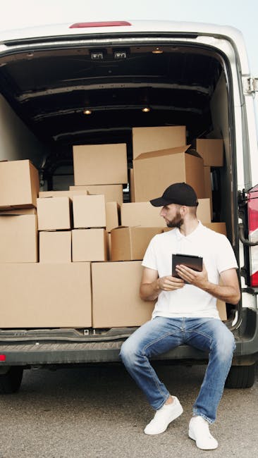 A man wearing a black cap, white t-shirt, and jeans is sitting on the pavement in front of a van with its rear doors open, revealing numerous brown cardboard boxes of various sizes packed tightly inside. The boxes are stacked on top of each other, some secured with plastic wrap, and are ready for home relocation. The man is holding a tablet device, possibly coordinating the packing and moving process as part of a professional house removal service. The setting suggests an outdoor loading area during daylight, with natural light illuminating the scene. Behind the van, a portion of the vehicle's interior is visible, emphasizing the loading process. This image supports services provided by Man with Van Maida Vale for end of tenancy removals, furniture transport, and packing and moving tasks related to property relocation.