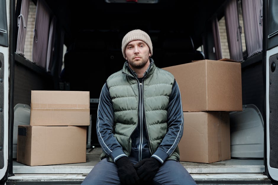 Two men wearing dark uniforms are loading a large, dark green sofa into the back of a white moving van parked on a paved street. The van's rear doors are open, revealing the interior with more packing materials and possibly other furniture. One man is holding the sofa steady while the other pushes it into the vehicle, both focused on the loading process. The background includes a lamppost, a street sign reading 'Somerset', and a bright, clear sky, indicating daytime. The scene depicts a professional home relocation or packing and moving service, with the movers carefully handling the furniture amidst an urban environment, supporting efficient furniture transport for house removals, as typical for end of tenancy relocations in Maida Vale. Man with Van Maida Vale may be involved in this process.
