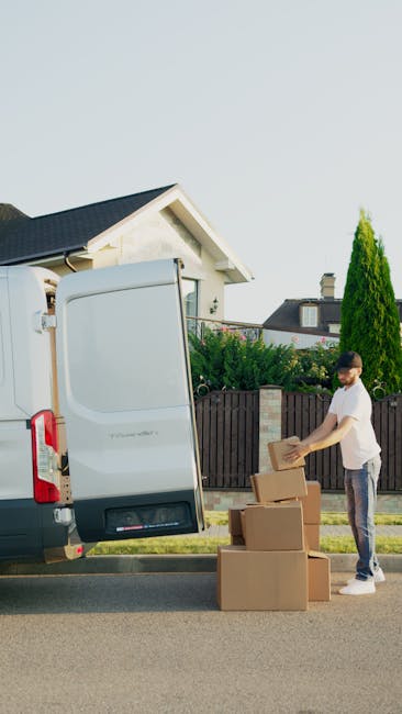 A young man with short dark hair, wearing a white t-shirt and grey trousers, is standing on a residential street during daylight hours, loading cardboard boxes into the rear of a white delivery van with its back doors open. The boxes are of various sizes, and some are stacked on the ground beside him. The van is parked near a pavement, with a wooden fence separating the property from the street. Behind the fence, there are green bushes and a tall, narrow conifer tree. In the background, part of a white house with a dark roof and a chimney, along with clear blue sky, is visible. The scene appears to be part of a house removal or moving process, involving packing and transporting household items, as managed by Man with Van Maida Vale, specialists in removals and furniture transport in the W9 area.