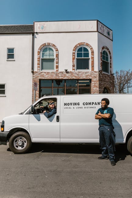 A white moving van with the branding 'MOVING COMPANY, LOCAL & LONG DISTANCE' parked in front of a two-storey residential building during daytime. The building features a white stucco and brick facade, with two large arched windows on the upper level and a row of smaller windows on the ground floor. The scene captures two movers engaged in a home relocation process: one inside the van appears to be communicating or giving instructions through the open driver’s side window, while the other stands outside, dressed in dark work clothes with arms crossed, observing the loading area. The pavement is clear, and there are visible equipment and materials such as cardboard boxes or packing supplies, suggesting the packing and furniture transport stages of a house move. Bright sunlight illuminates the scene, with a clear blue sky overhead. This image supports house removals services by illustrating the loading process involved in home relocation, as provided by Man with Van Maida Vale.