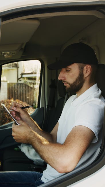 A young man with short dark hair, wearing a white t-shirt and grey trousers, is standing on a residential street during daylight hours, loading cardboard boxes into the rear of a white delivery van with its back doors open. The boxes are of various sizes, and some are stacked on the ground beside him. The van is parked near a pavement, with a wooden fence separating the property from the street. Behind the fence, there are green bushes and a tall, narrow conifer tree. In the background, part of a white house with a dark roof and a chimney, along with clear blue sky, is visible. The scene appears to be part of a house removal or moving process, involving packing and transporting household items, as managed by Man with Van Maida Vale, specialists in removals and furniture transport in the W9 area.