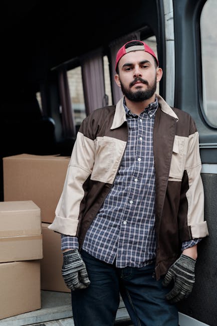 A man with a beard and dark hair, wearing a red baseball cap backwards, a plaid shirt, and a beige and brown work jacket, stands inside the rear of a moving van. To his left are stacked cardboard boxes of various sizes, some wrapped in clear plastic and others open, revealing packing materials. The van's interior features dark panels and windows, with the scene illuminated by natural light. The man appears to be involved in home relocation, possibly overseeing or preparing for furniture transport and packing for a house move undertaken by Man with Van Maida Vale. The environment suggests an organized loading process with objects arranged for easy handling during a house removals service, supporting efficient home relocation logistics.