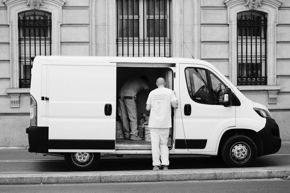 A white moving van with its side door open, parked on a street in front of an ornate stone building with barred windows. Inside the van, several individuals are engaged in a home relocation process, handling various packing materials including cardboard boxes, blankets, and plastic wrapping. One person is bending over, possibly organizing or lifting items, while another stands outside the van, observing or assisting. The scene depicts the loading or unloading phase of furniture transport and packing during a professional removal service, with clear identification of the vehicle, packaging materials, and team members involved in the furniture transport operation. The lighting appears natural, highlighting the details of the moving equipment and the urban environment, aligning with standard house removals and relocation activities carried out by Man with Van Maida Vale.