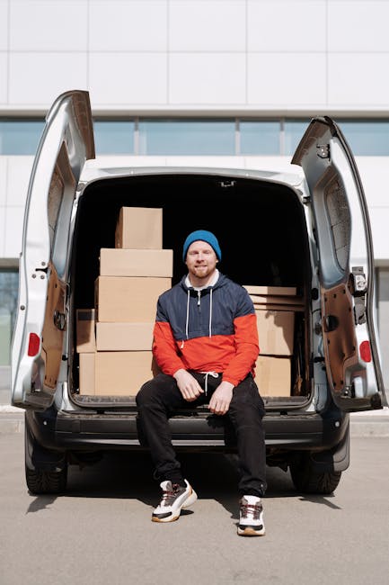 A man with a beard and dark hair, wearing a red baseball cap backwards, a plaid shirt, and a beige and brown work jacket, stands inside the rear of a moving van. To his left are stacked cardboard boxes of various sizes, some wrapped in clear plastic and others open, revealing packing materials. The van's interior features dark panels and windows, with the scene illuminated by natural light. The man appears to be involved in home relocation, possibly overseeing or preparing for furniture transport and packing for a house move undertaken by Man with Van Maida Vale. The environment suggests an organized loading process with objects arranged for easy handling during a house removals service, supporting efficient home relocation logistics.